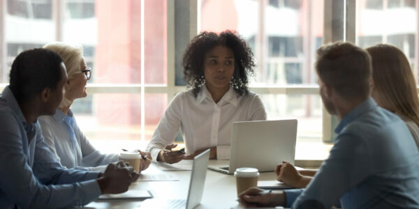 Five multi-ethnic businessman businesswoman gathered together at modern boardroom for planning future projects discussing relevant topics. Black team leader or coach sitting at desk talking with staff